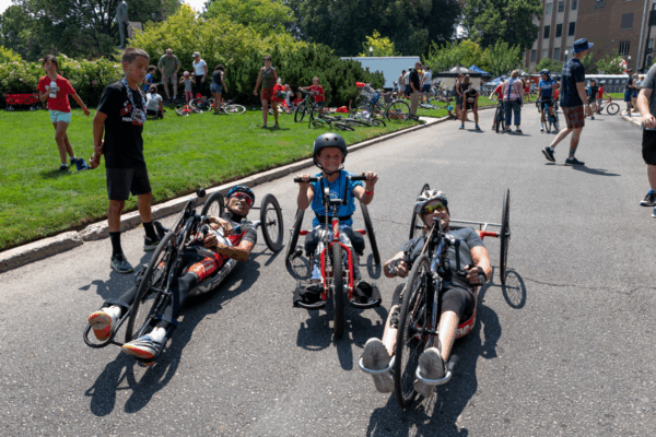 Two adult handcyclists and a young child on a smaller red handcycle smile together on the road, surrounded by families and other cyclists during an inclusive community event.