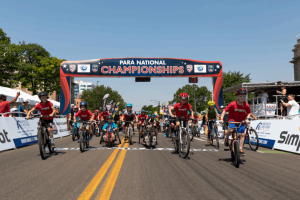 A group of children and youth riders, including a young handcyclist, start under a “Para National Championships” archway on a sunny day, celebrating inclusive cycling competition.
