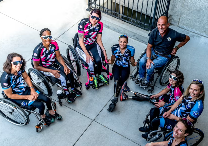 A group of CAF athletes, several using wheelchairs and prosthetics, smile together in a circle with a coach, all wearing matching cycling team kits.