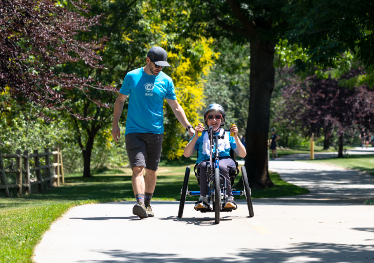 CAF athlete assists a young rider using a handcycle on a sunny park path.