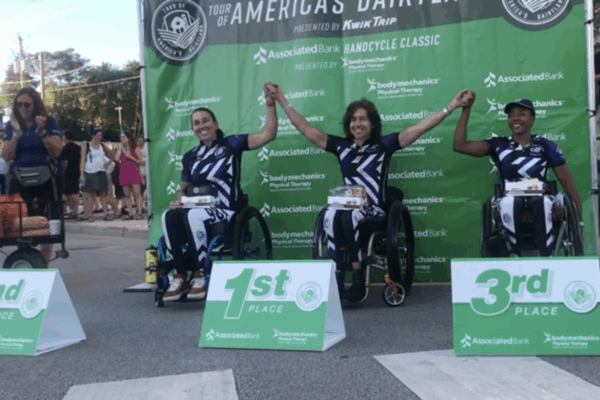 Three CAF women’s handcyclists celebrate on the podium, raising their hands after winning first, second, and third place.