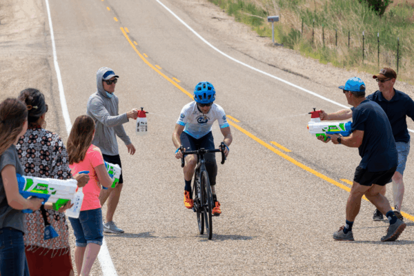 A cyclist wearing a CAF jersey and blue helmet rides uphill as supporters on both sides of the road spray them with water and cheer in the summer heat.
