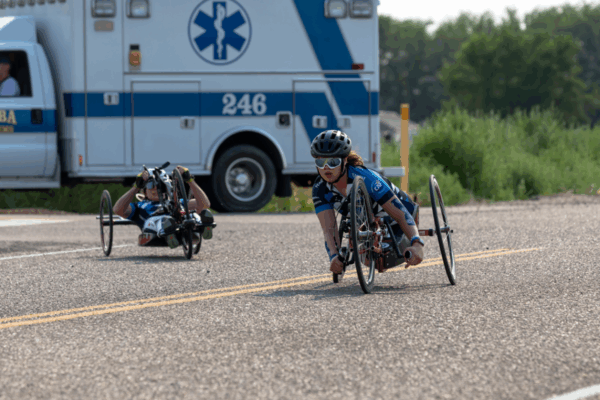 Two athletes with physical disabilities race along the road in handcycles, wearing helmets and blue CAF team jerseys, with an ambulance parked in the background for event safety.