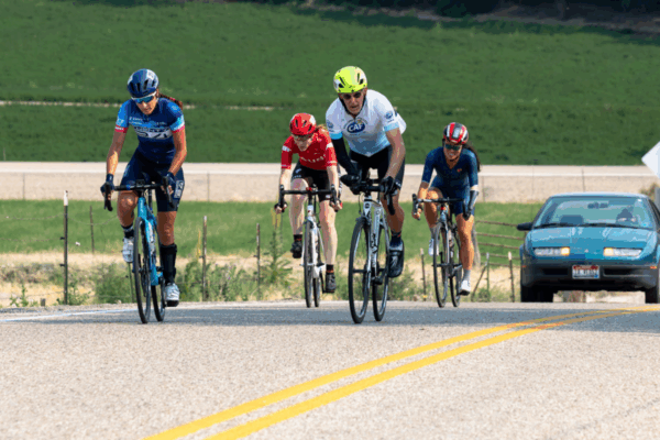 Four cyclists, including one wearing a CAF jersey and bright yellow helmet, climb a hill during a road race. The riders are focused and determined as a car follows behind them on the course.