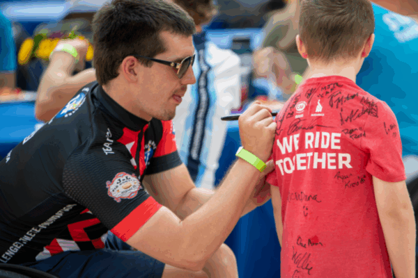 An athlete with a physical disability wearing a CAF cycling jersey signs the back of a young boy’s red shirt that reads “WE RIDE TOGETHER,” surrounded by other participants and families at the event.
