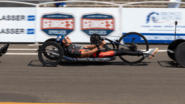 A male athlete races in a handcycle on a paved road, wearing a helmet and sunglasses, with “Paralyzed Veterans of America” printed on the side of his racing chair.