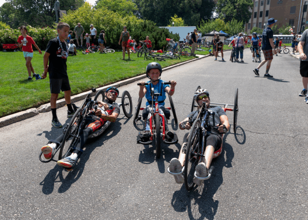 Two adult athletes in handcycles smile alongside a young child riding a smaller adaptive cycle, surrounded by other participants and families at an outdoor cycling event.