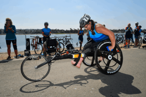 Young athletes using racing wheelchairs and everyday wheelchairs gather on a track with coaches, preparing for training, with mountains in the background.