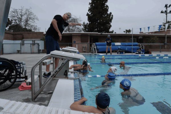 A coach stands on the pool deck giving instructions to a group of youth athletes in swim caps and goggles, who are gathered in the water at swim practice.