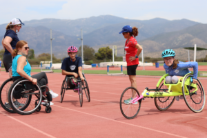 Kids in wheelchair racing chairs on track next to Mary Kate Callahan and coaches