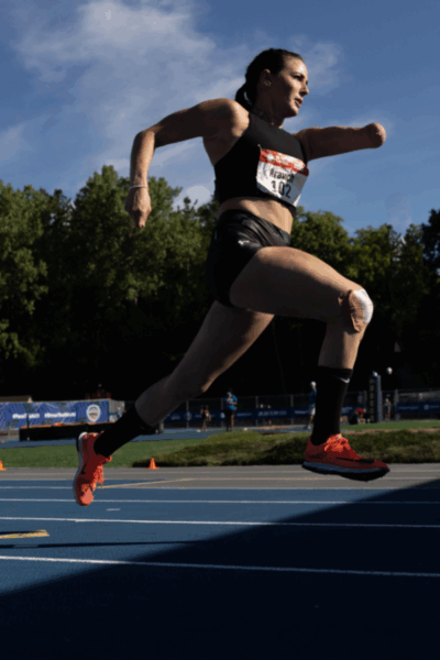 A female sprinter with a limb difference powers down the track in competition, her muscles tensed and orange racing shoes striking the blue surface.