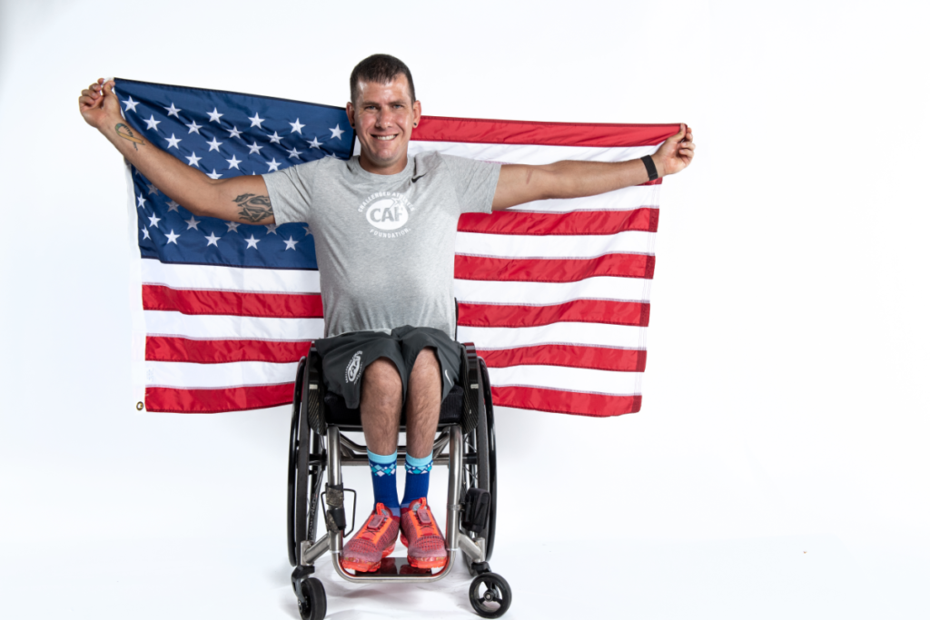 Erik Hightower in grey CAF shirt sitting in wheelchair with American flag held behind his head