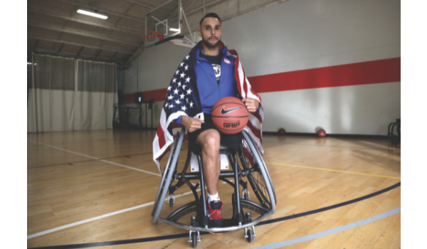 Jorge Sanchez sitting in basketball wheelchair with American flag draped around his shoulders and basketball in lap