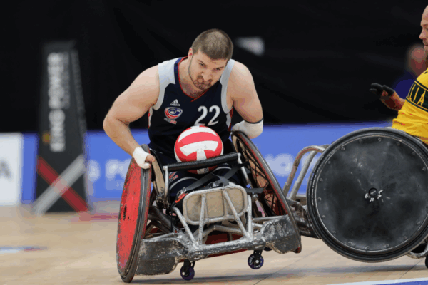 A male wheelchair rugby athlete wearing jersey number 22 grips the ball tightly while maneuvering his chair on the court during a game.