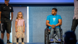 Young man in wheelchair with CAF Idaho shirt on and young girl standing together on stage