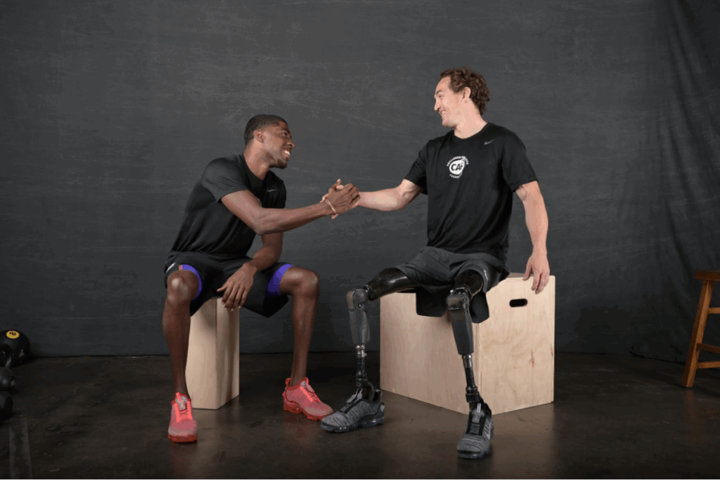 Two athletes sit on wooden boxes, smiling and shaking hands during a photo shoot.