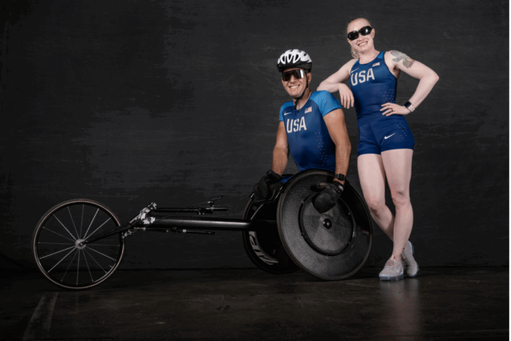 Two Team USA athletes pose in blue racing uniforms, one standing and one in a racing wheelchair, against a dark studio backdrop.