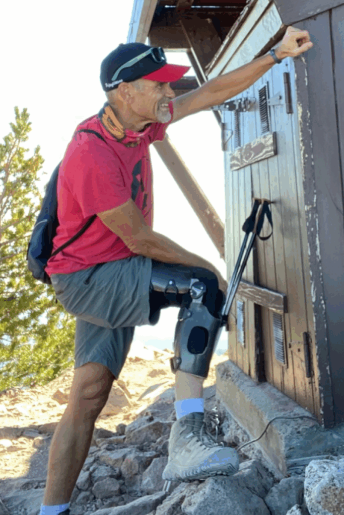 Smiling hiker wearing a red shirt and an orthotic leg brace leans against a wooden structure on a rocky trail.