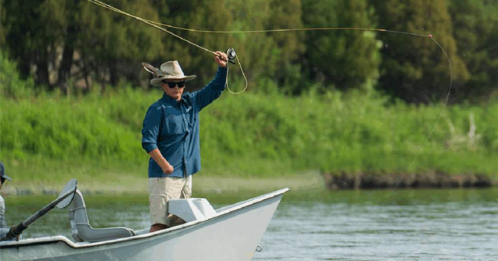 A camper stands on a drift boat casting a fishing line across the river on a sunny day, with trees lining the riverbank.