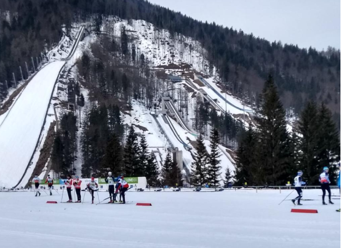Athletes prepare on a snow-covered Nordic ski course at the Slovenia World Cup, with towering ski jumps and forested mountains rising behind them.
