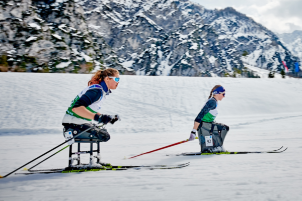 Two athletes with physical disabilities race side by side in Nordic sit-skiing, using poles to propel themselves across a snowy course with rocky mountain peaks behind them.
