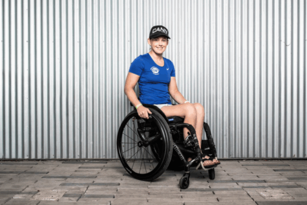 A woman in a blue CAF shirt and black hat smiles at the camera while seated in a wheelchair in front of a corrugated metal wall.