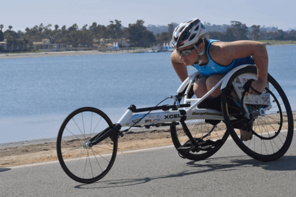 An athlete races along a waterfront road in a white racing wheelchair, wearing a helmet and sunglasses, with the ocean and palm trees visible in the background.