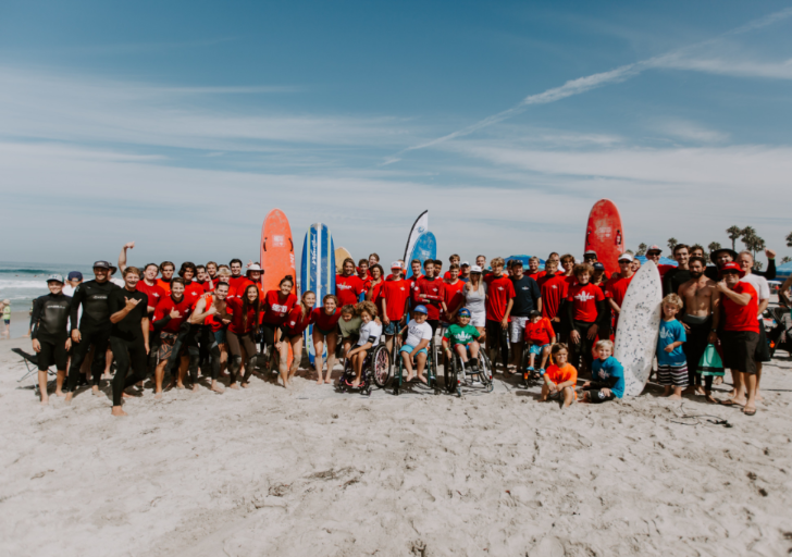 A large group of athletes, volunteers, and families gather on the beach in front of surfboards, many wearing red shirts, smiling together for a group photo.