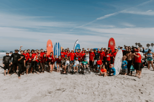A large group of athletes, volunteers, and families gather on the beach in front of surfboards, many wearing red shirts, smiling together for a group photo.