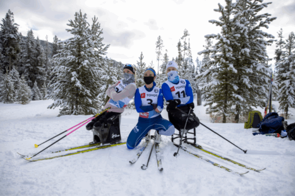 Three athletes in sit-skis wearing Toyota racing bibs pose playfully in the snow among tall pine trees, smiling and striking fun poses with their ski poles.