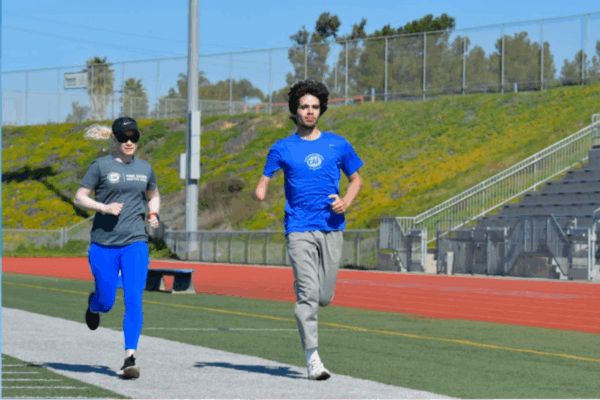 Two athletes run side by side on a track during a CAF training session, one wearing a gray shirt and bright blue pants, the other in a blue CAF shirt with a visible limb difference.