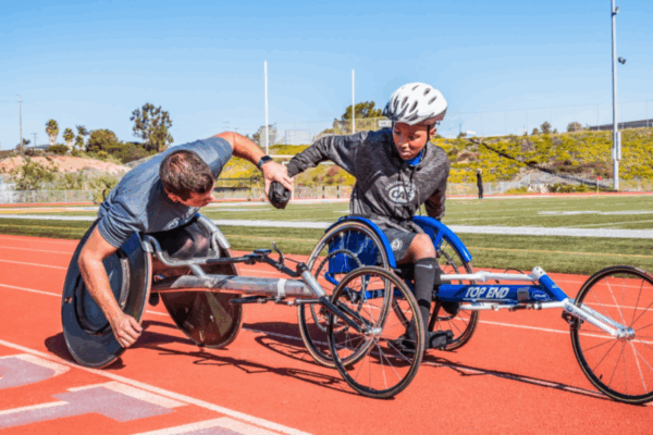 A coach leans down to guide a young athlete in a racing wheelchair on a track. Both wear CAF shirts, and the athlete looks focused as they prepare to push forward.