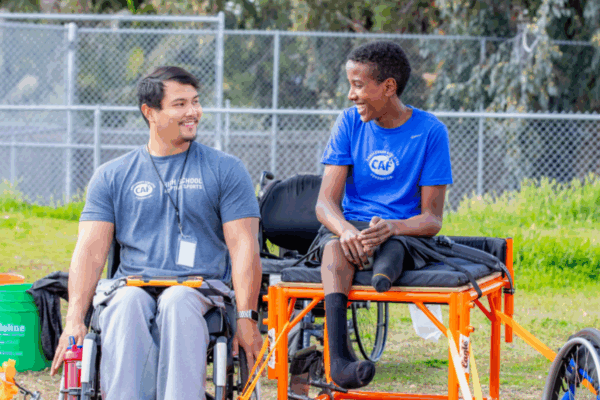 Two athletes in wheelchairs, one adult and one youth, sit side by side smiling at each other during a CAF High School Adaptive Sports program practice.