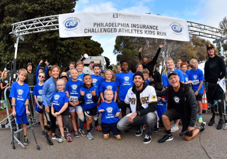 A large group of children in bright blue CAF shirts gather at the starting line under a “Challenged Athletes Kids Run” banner. Several kids with prosthetics and mobility aids stand proudly among their peers, joined by adults and volunteers, all smiling at the camera.
