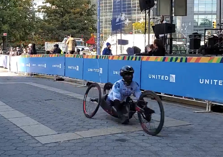 An athlete with a physical disability wearing a helmet and racing jersey pushes forward in a handcycle near the finish line, with spectators clapping behind barricades.