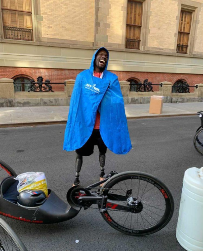 The athlete stands with prosthetic running blades, laughing while draped in a blue NYC Marathon finisher’s poncho, next to his handcycle.
