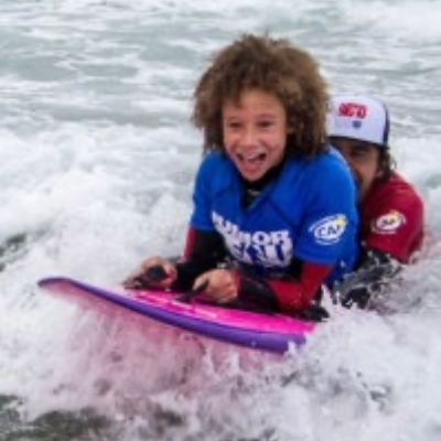 A child with a physical disability wearing a blue CAF Junior Team shirt rides a pink bodyboard on a wave, smiling with excitement as a surf instructor supports from behind.