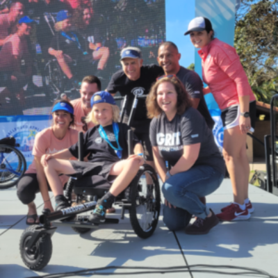 A young athlete with a physical disability sits in an adaptive handcycle on stage, surrounded by smiling adults and teammates posing together for a photo.