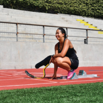 A female athlete with a physical disability, using a running blade prosthetic, crouches at the starting blocks on a track while smiling during training.