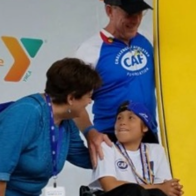 A child athlete with a physical disability wearing a CAF shirt and cap smiles up at an adult leaning toward him, while another adult in a CAF jersey stands nearby on stage.