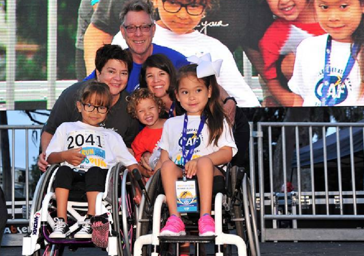 Two young athletes with physical disabilities sit in wheelchairs wearing medals and CAF shirts, joined by friends and adults smiling proudly on stage.