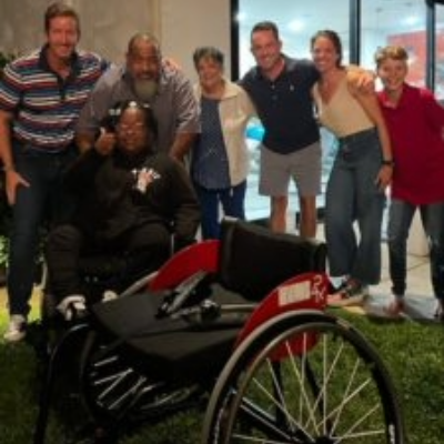 A group of six people stand behind an athlete with a physical disability seated in a racing wheelchair, all smiling and posing together outdoors at night.