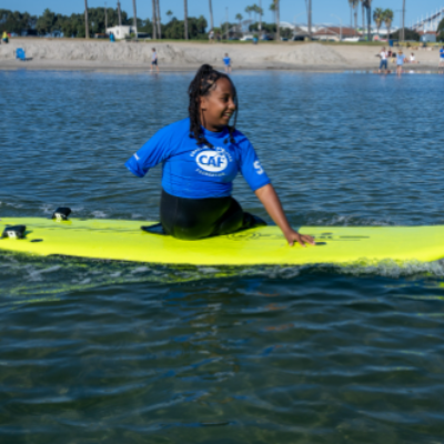 A young athlete with a physical disability wearing a blue CAF shirt balances on a bright yellow surfboard in calm ocean water.