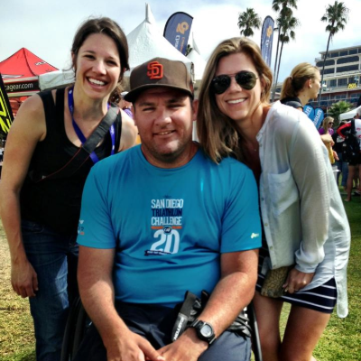 An athlete with a physical disability wearing a blue San Diego Triathlon Challenge shirt sits in a wheelchair, smiling with two women standing beside him at an outdoor event.