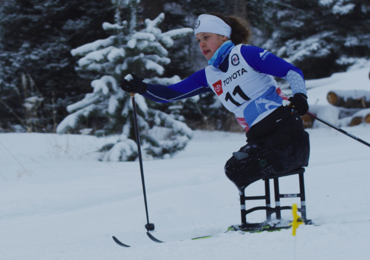 Athlete with a physical disability competes in an adaptive Nordic sit-ski race, using ski poles to propel forward on a snowy course surrounded by evergreen trees.