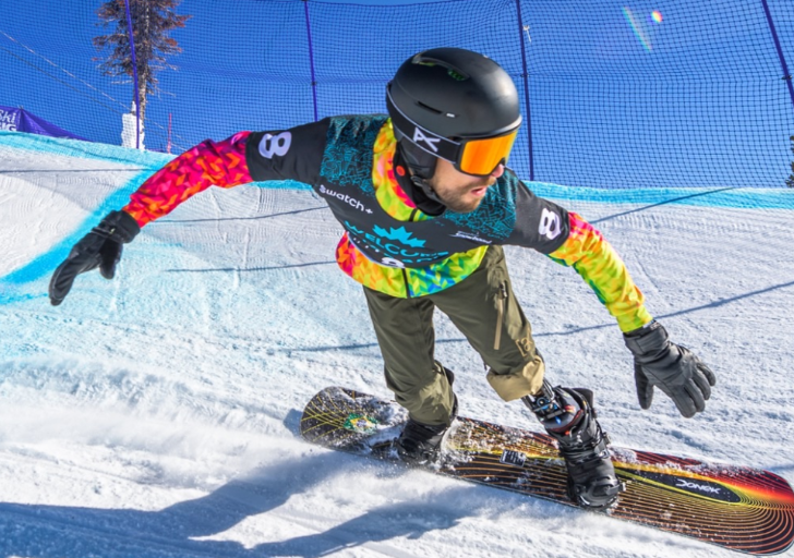 Snowboarder with a physical disability carves through a banked turn on a snowy course during a competition, wearing a helmet and goggles.
