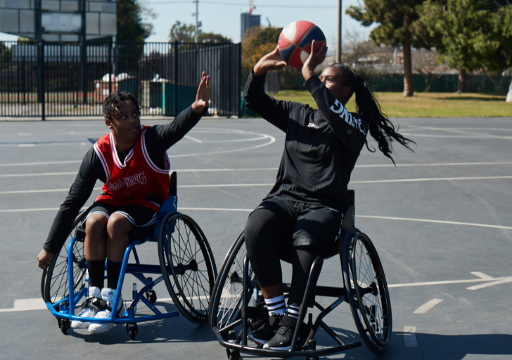 Two athletes with physical disabilities compete in wheelchair basketball on an outdoor court as one player shoots while another defends.