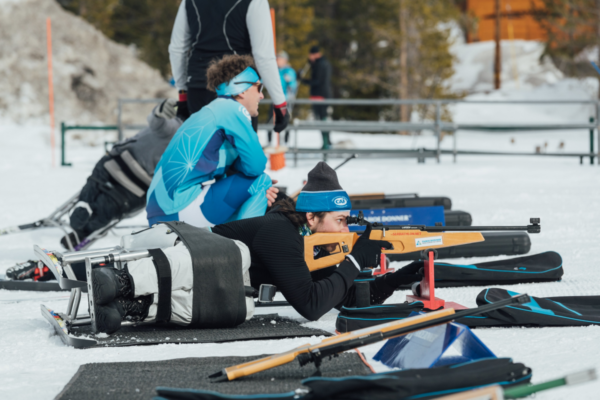 An athlete with a physical disability lies prone on the snow aiming a biathlon rifle during an adaptive Nordic ski and biathlon clinic, while another participant and coach observe nearby.