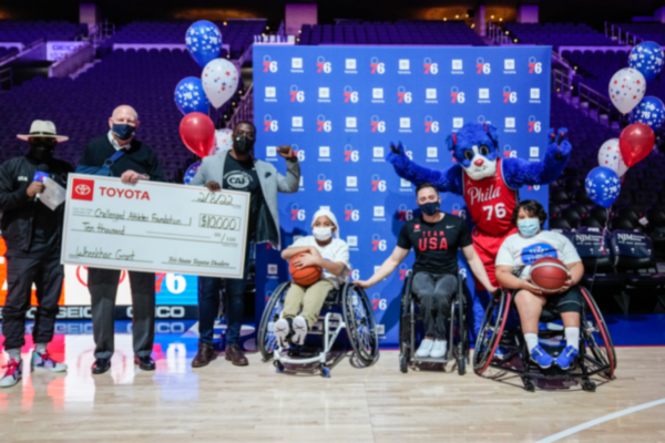 CAF youth athletes with physical disabilities sit on a basketball court alongside partners and supporters during a check presentation event, celebrating community support for adaptive sport.