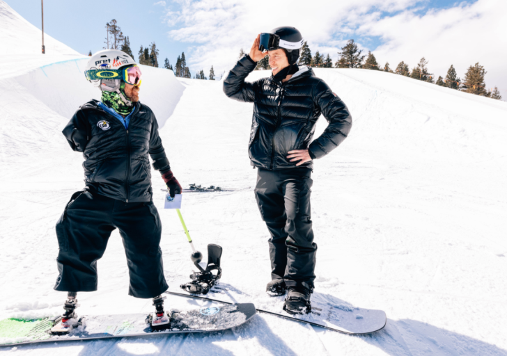 Two snowboarders, one with prosthetic legs, pause on a sunny slope to talk during a winter sports session.
